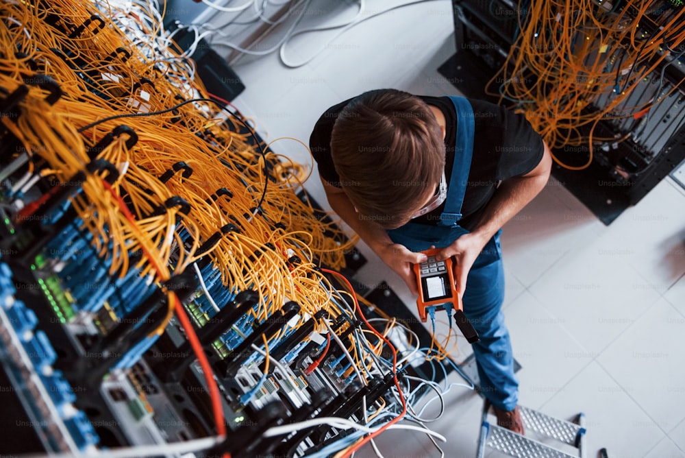 Vista dall'alto del giovane in uniforme con dispositivo di misurazione che funziona con apparecchiature e cavi internet nella sala server.