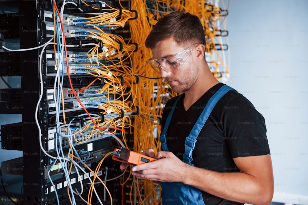 Young man in uniform with measuring device works with internet equipment and wires in server room.