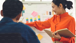 Young woman explains business data on white board in casual office room . The confident Asian businesswoman reports information progress of a business project to partner to determine market strategy .