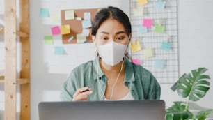Asia businesswoman wearing medical face mask using laptop talk to colleagues about plan in video call while working from home at living room. Social distancing, quarantine for corona virus prevention.