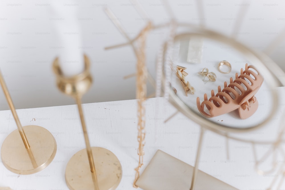 Des accessoires dorés modernes et des pinces à cheveux se reflètent dans un miroir bohème sur une table blanche avec des bougies vintage. Bague en or élégante, collier chaîne, boucles d’oreilles, épingles à cheveux et barrettes. Bijoux bohèmes