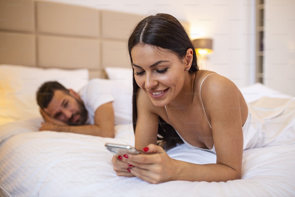 young couple in bed husband frustrated upset and unsatisfied while his internet addict wife is using mobile phone in social network addiction ignoring him in relationship domestic problems