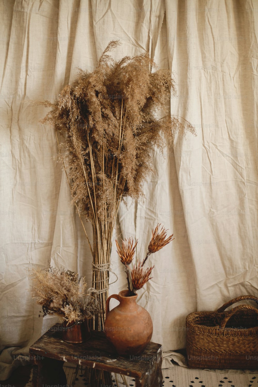 Dry protea flowers in clay vase, pampas grass, wicker basket on boho rug on background of beige linen cloth. Atmospheric bohemian style room details. Boho decor for wedding or in studio