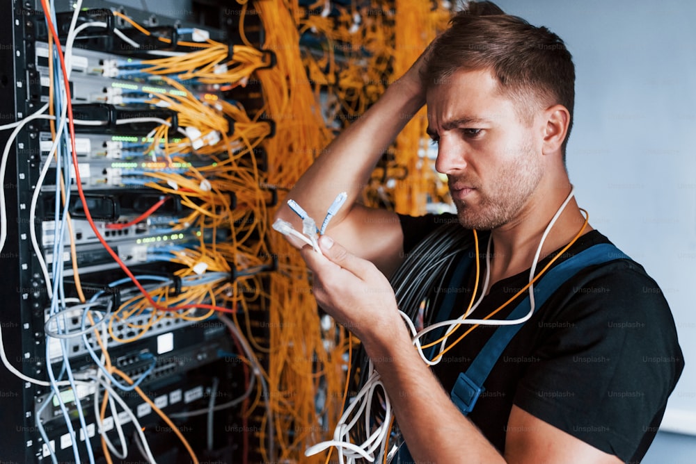 Young man in uniform feels confused and looking for a solution with internet equipment and wires in server room.