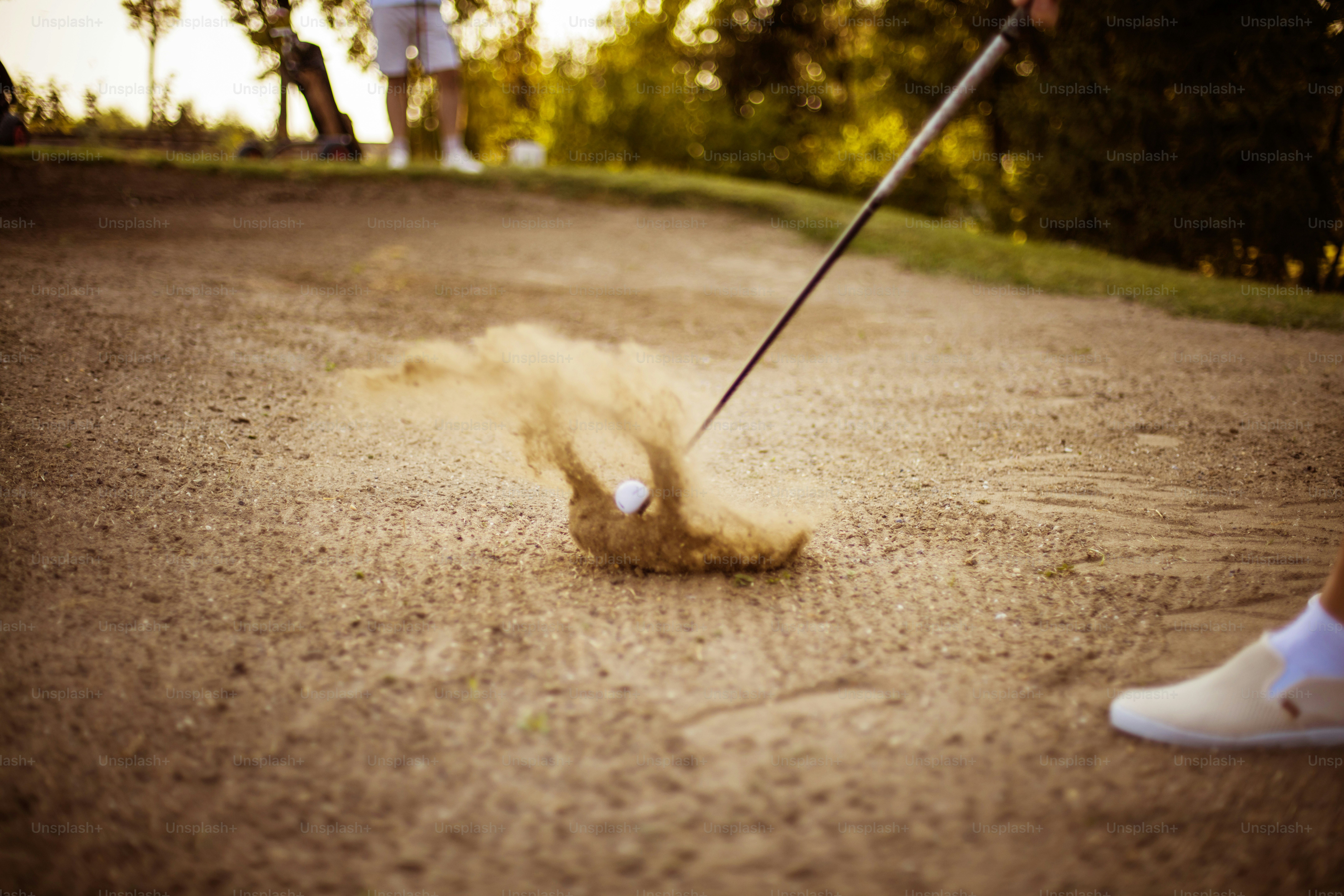 Two men on the golf course. Man hits a golf ball. Focus is on sand.