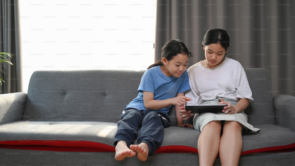 Dos hermanas sentadas en el sofá y viendo dibujos animados en línea con una tableta digital en casa.