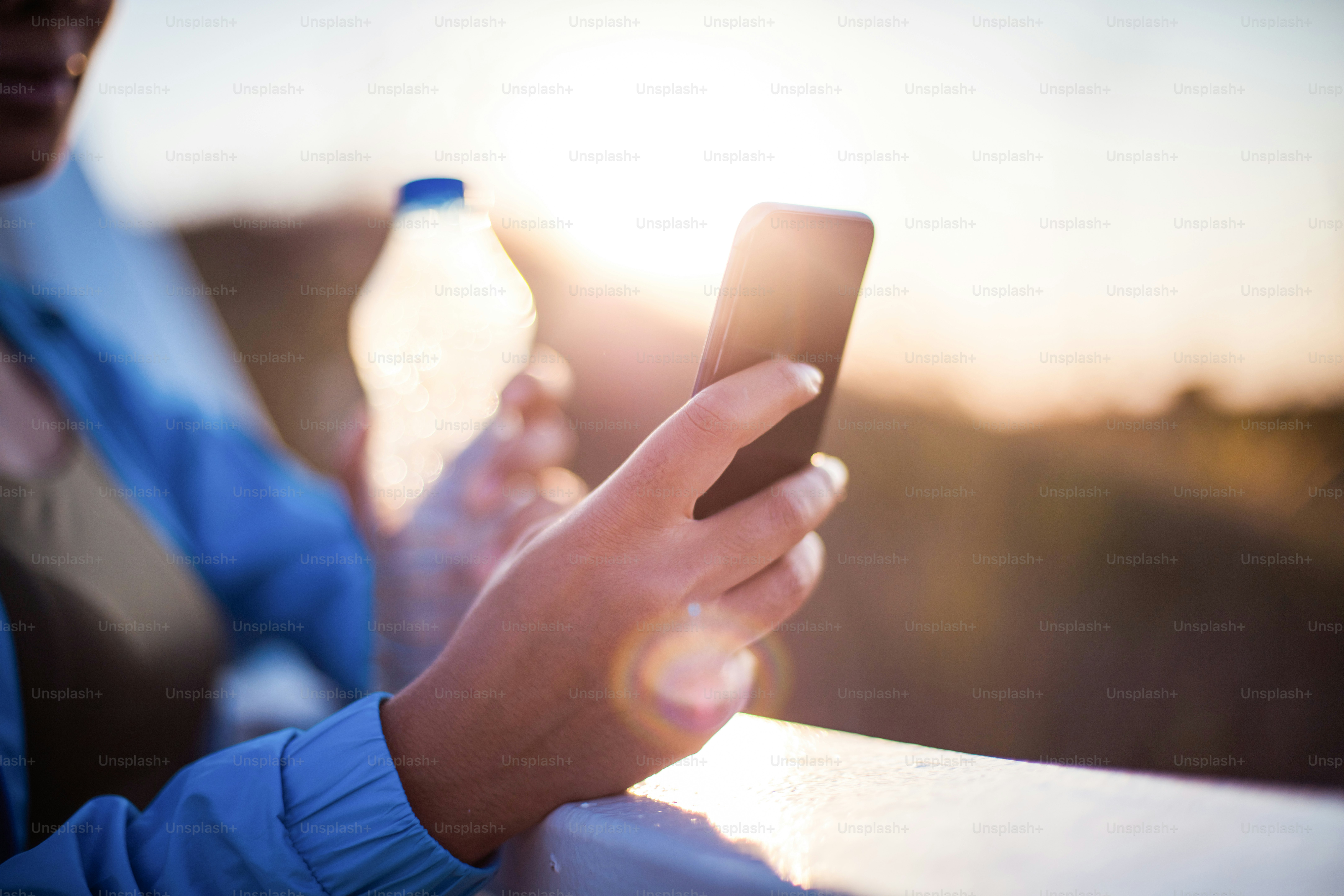 African sports woman standing in nature, holding bottle of water and using smart phone. Focus is on hand.
