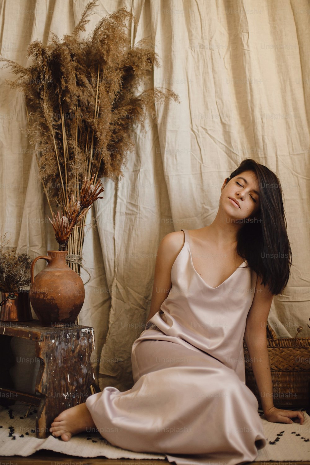 Stylish fashionable woman in silk dress sensually posing in boho style room with dry grass and flowers on beige background. Modern bohemian bride. Young attractive female in atmospheric interior