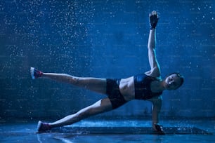 Muscular wet young woman training on floor on mat in empty hall under rain, dark blue light. Female bodybuilder in gloves practicing side plank exercise with hand and leg up indoors. Concept of sport.