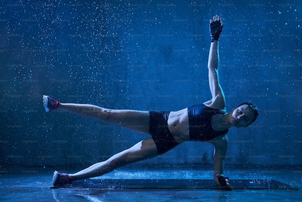 Muscular wet young woman training on floor on mat in empty hall under rain, dark blue light. Female bodybuilder in gloves practicing side plank exercise with hand and leg up indoors. Concept of sport.