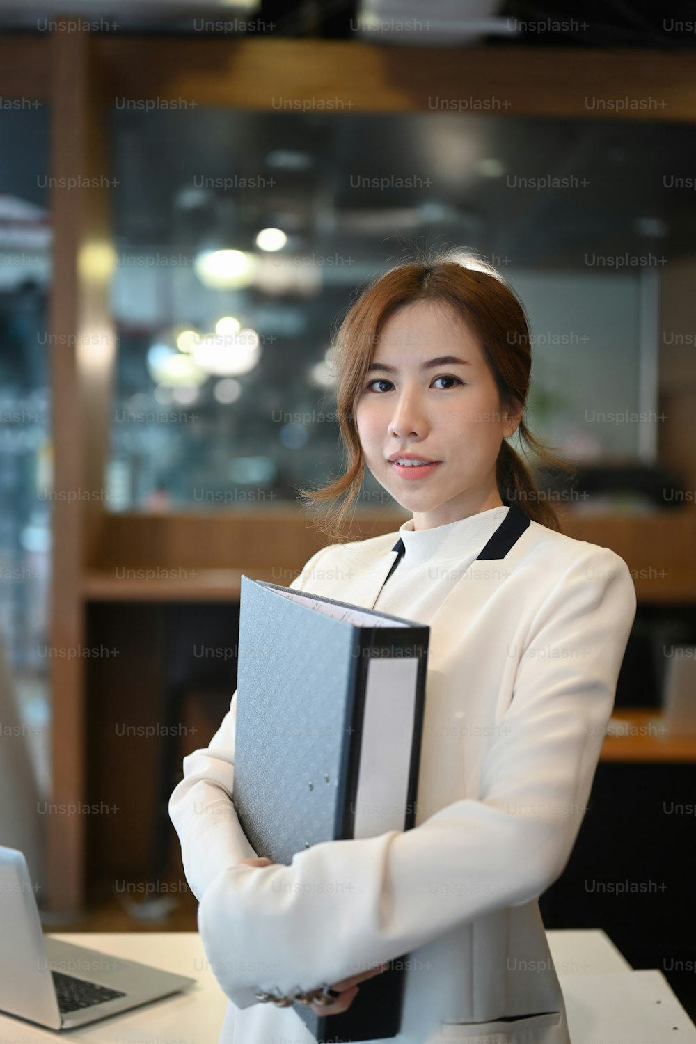 Porträt einer selbstbewussten Geschäftsfrau, die eine Geschäftsmappe hält und in einem modernen Büro steht.