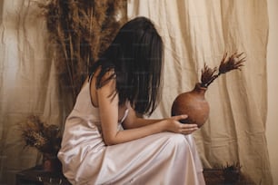 Mujer sensual con estilo en vestido de seda sosteniendo jarrón de arcilla con flores en habitación de estilo bohemio con hierba seca. Joven mujer de moda posando en interior boho atmosférico. Novia moderna