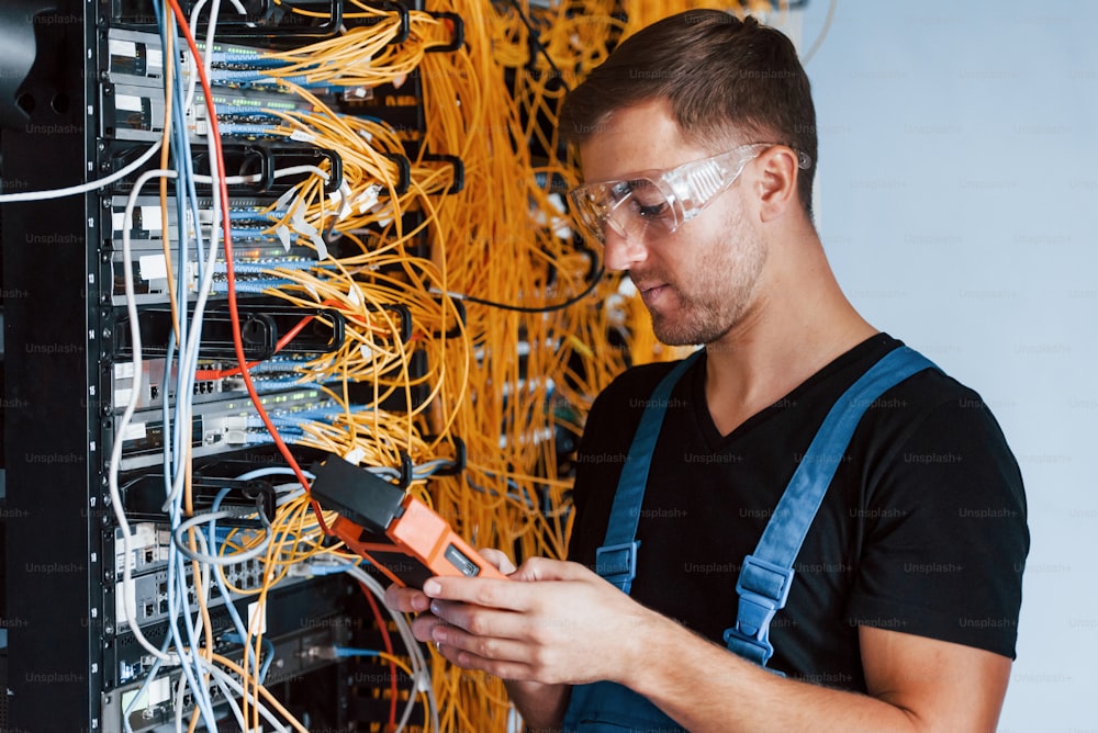Young man in protective glasses works with internet equipment and wires in server room.
