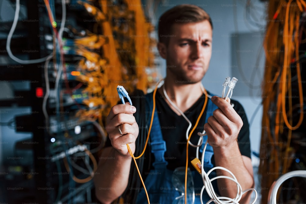 Young man in uniform feels confused and looking for a solution with internet equipment and wires in server room.