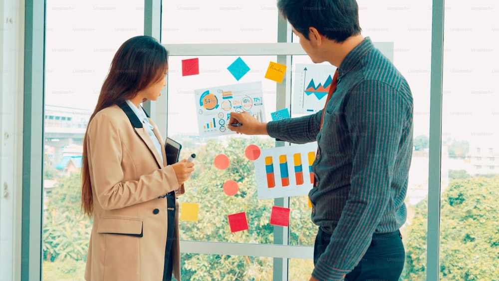 ビジネスマンは、オフィスでプロジェクト計画ボードに取り組み、同僚の友人と会話してプロジェクト開発を分析します。ガラスの壁に貼られた付箋を使って整理整頓します。