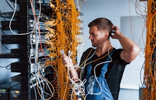 Young man in uniform feels confused and looking for a solution with internet equipment and wires in server room.