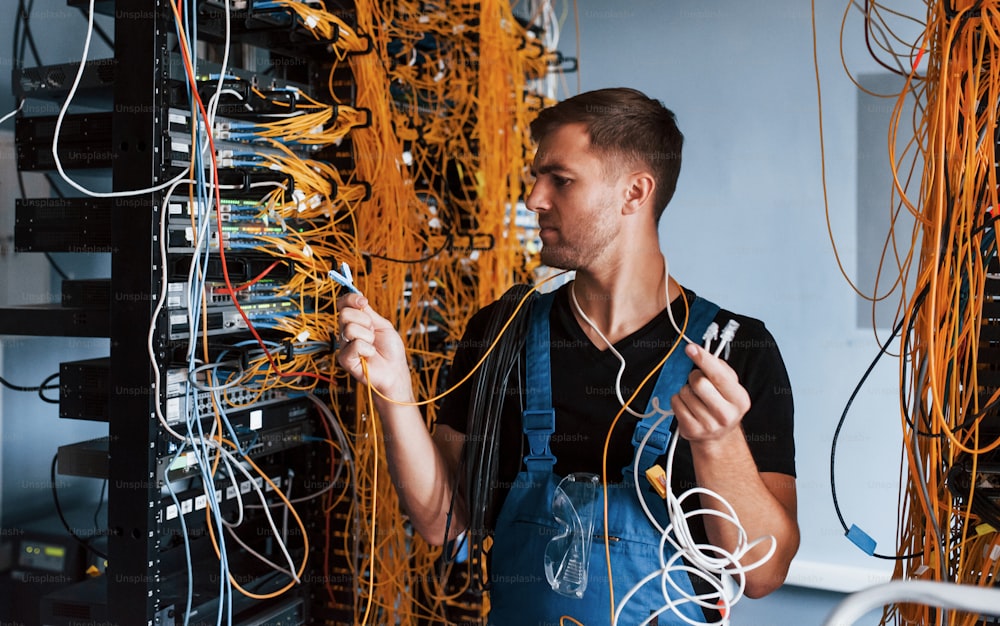 Young man in uniform feels confused and looking for a solution with internet equipment and wires in server room.