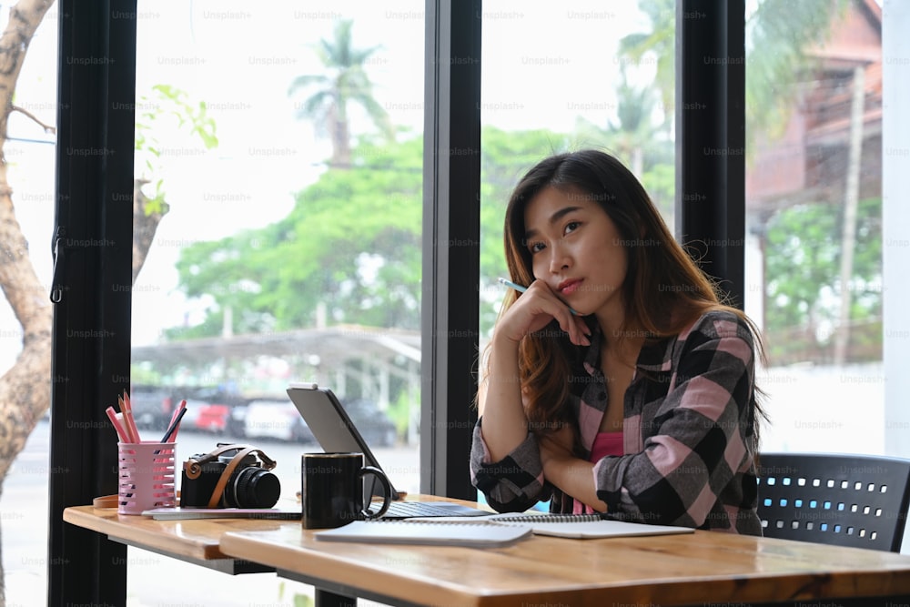 Pensive young woman designer looking away thinking future project while sitting at her workplace.