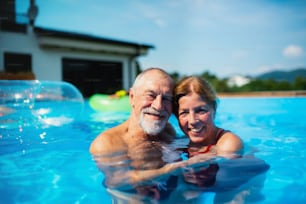 Portrait of cheerful senior couple in swimming pool outdoors in backyard, looking at camera.