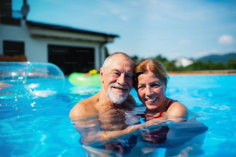 Retrato do casal de idosos alegres na piscina ao ar livre no quintal, olhando para a câmera.