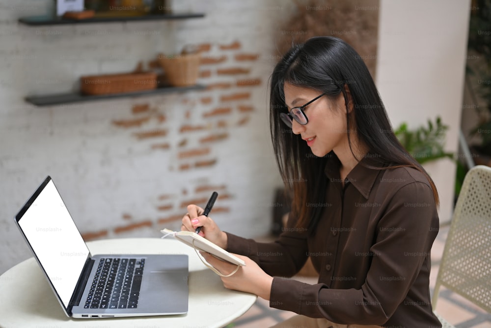 Vista lateral de una mujer joven que trabaja con el libro de horarios y la computadora portátil en la mesa redonda en el café