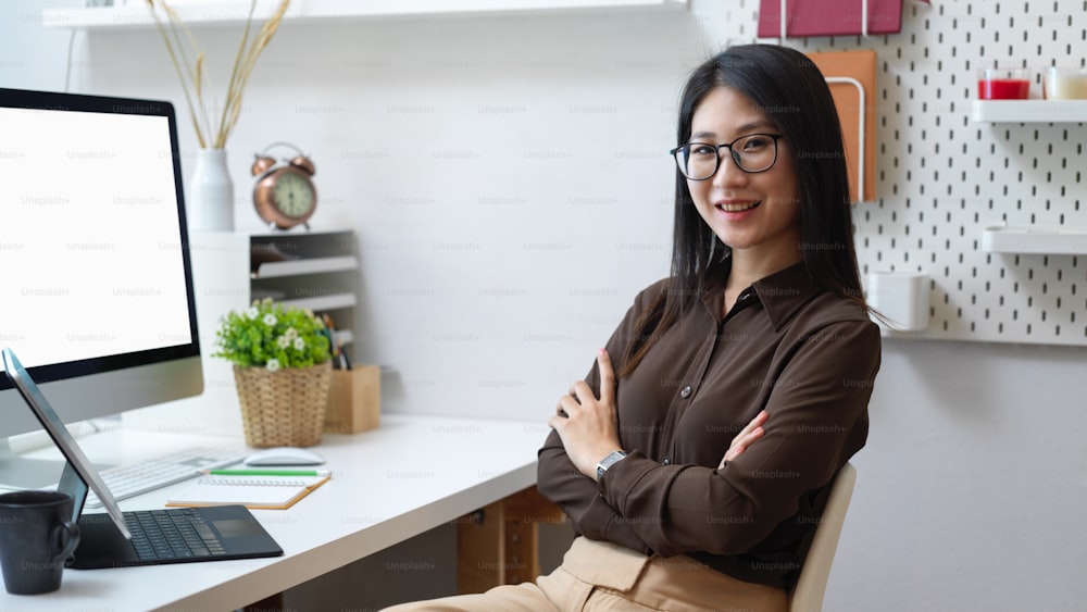 Young female office worker with eyeglasses smiling to camera while sitting in home office room