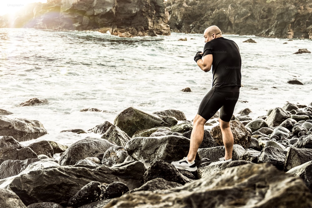 Photo of a strong sports man doing boxer training outdoors at the rocky beautiful beach . Healthy lifestyle and sport concept.