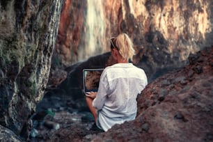 Woman working with computer or talking with family and friends online, sitting on the wild place in the rocky muntains. Remote work concept during covid pandemia.