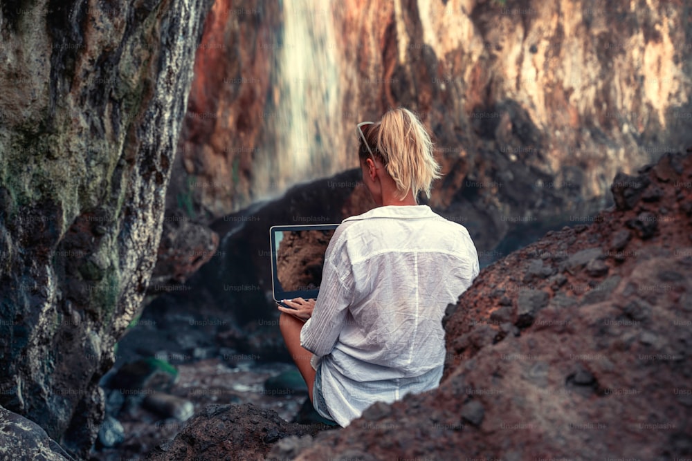 Woman working with computer or talking with family and friends online, sitting on the wild place in the rocky muntains. Remote work concept during covid pandemia.