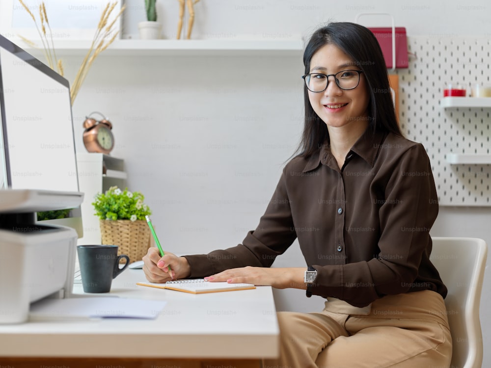 Side view of young female office worker in eyeglasses smiling to camera while sitting at office desk