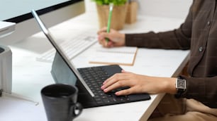 Vista lateral da mão feminina que trabalha com tablet digital e escreve no caderno na mesa do computador