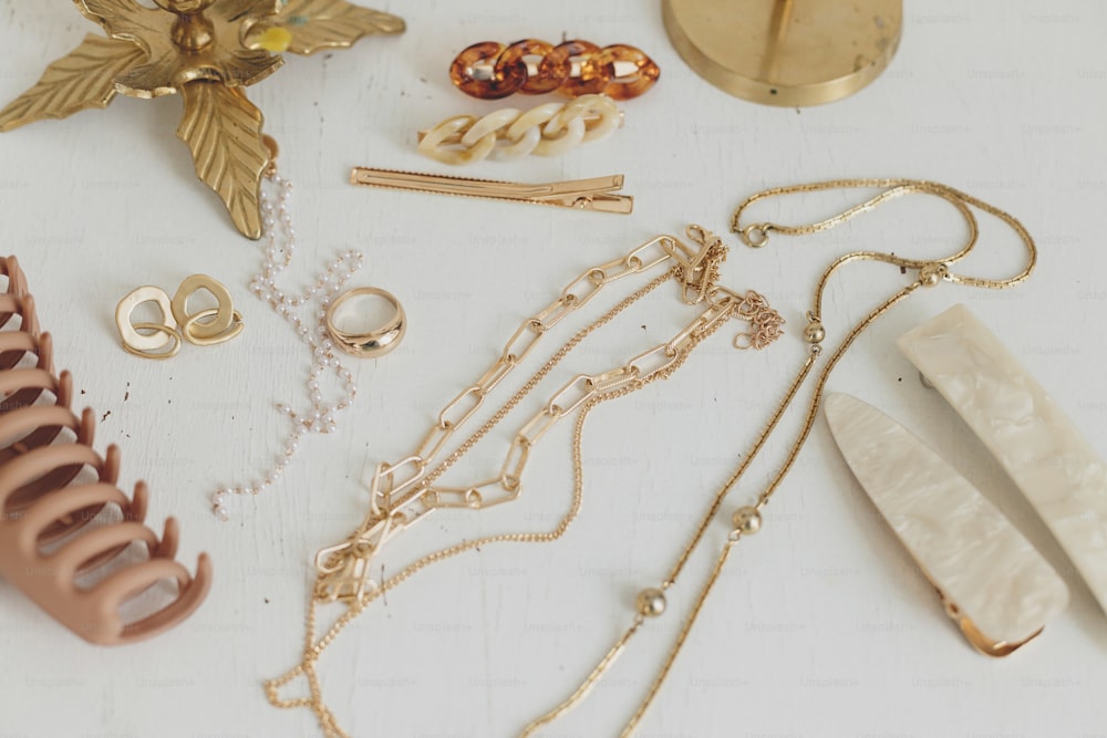 Joyas doradas modernas y pinzas para el cabello en mesa de madera blanca con candelabros vintage. Elegante anillo de oro, collar de cadena, pendientes, horquillas y pasadores beige. Accesorios boho