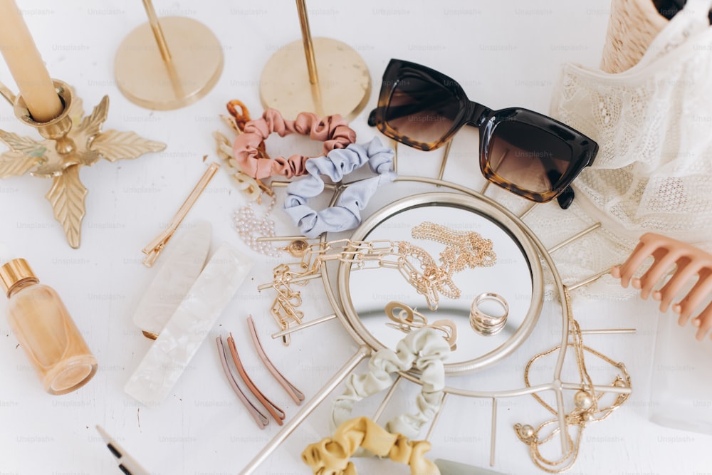 Modern summer accessories. Golden jewellery, sunglasses, hair clips, cosmetics, perfume and lace lingerie on white table with vintage candles and boho mirror. Feminine essentials