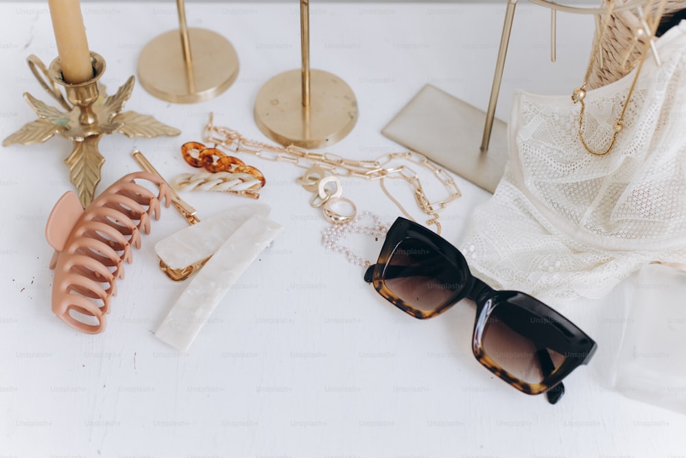 Golden jewellery, sunglasses, hair clips, cosmetics, perfume and lace lingerie on white table with vintage candles and boho mirror. Modern summer accessories. Feminine essentials