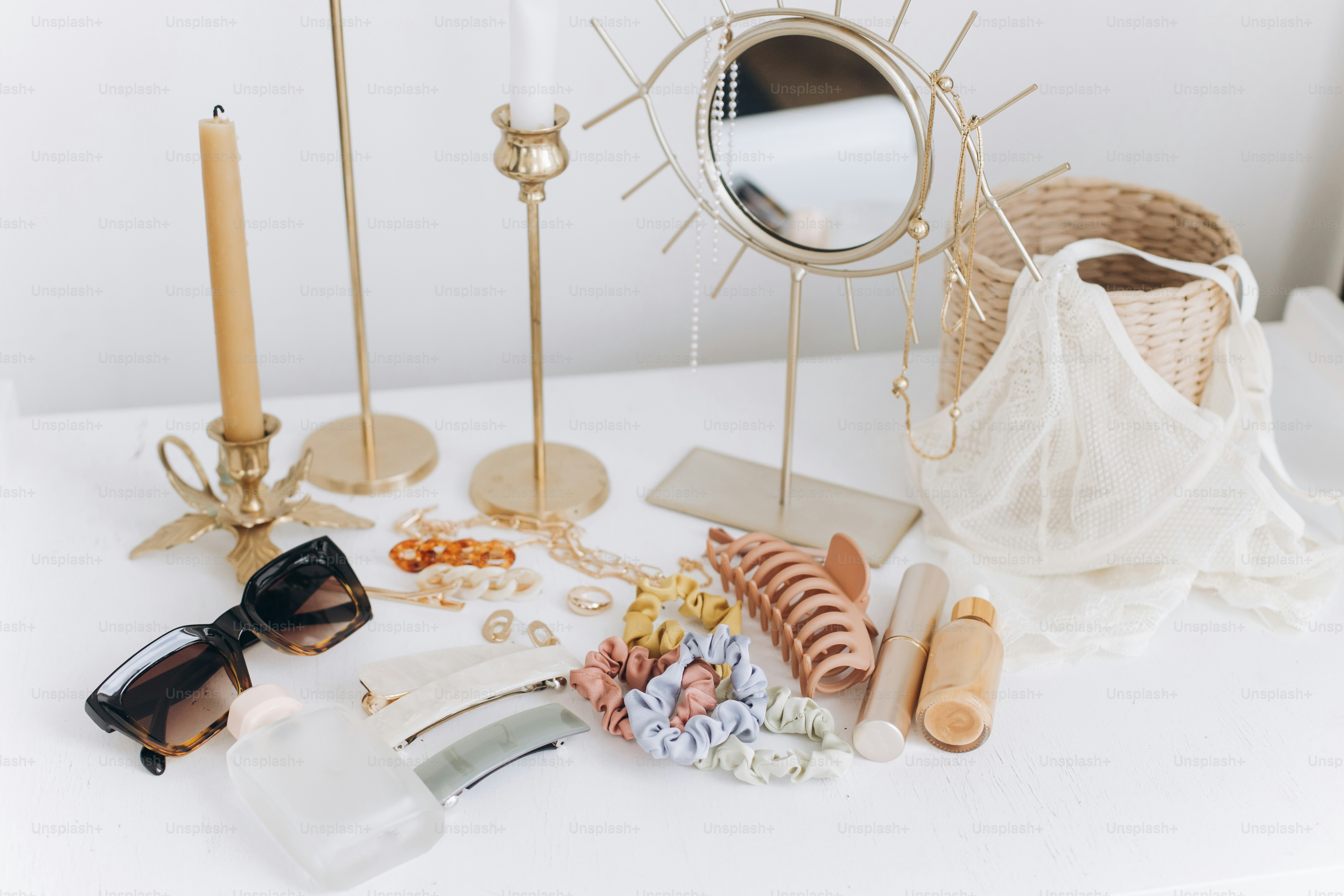 Modern summer accessories. Golden jewellery, sunglasses, hair clips, cosmetics, perfume and lace lingerie on white table with vintage candles and boho mirror. Feminine essentials