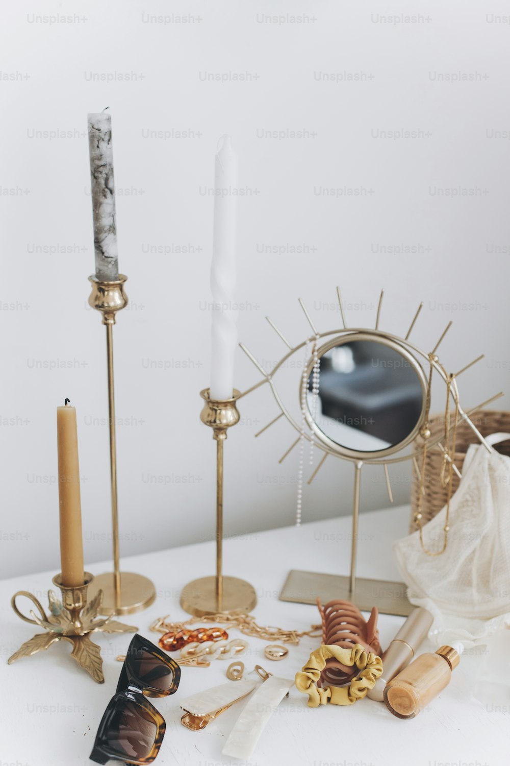 현대 여름 액세서리. 황금 보석, 선글라스, 헤어 클립, 화장품, 향수 및 레이스 란제리가 빈티지 양초와 보헤미안 거울이있는 흰색 테이블에 있습니다. 여성스러운 필수품