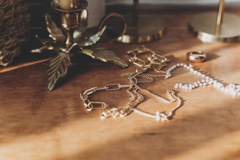 Moderne Gold- und Perlenaccessoires auf dem Tisch mit Vintage-Kerzenhaltern. Stilvolle goldene Halskette und Ring auf Holzhintergrund in sonnigem Licht.