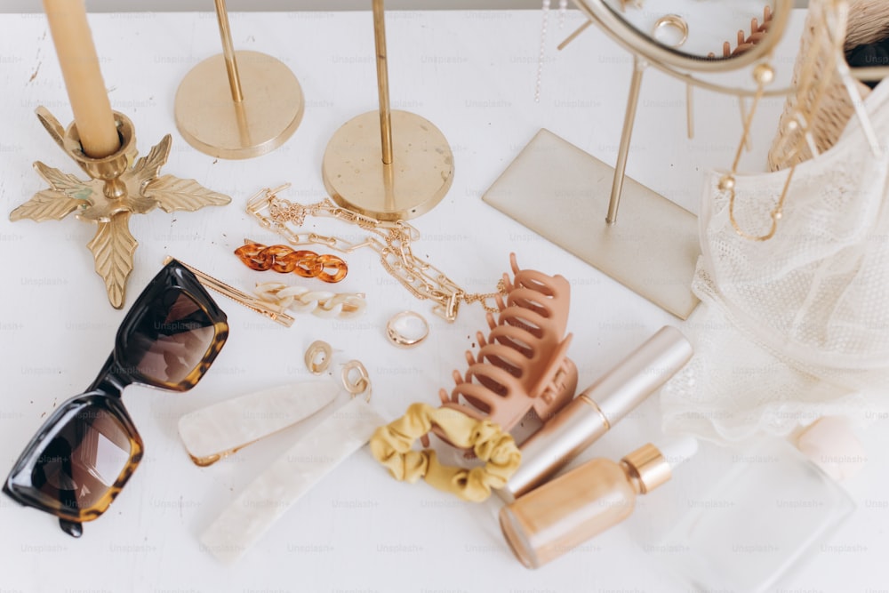 Modern summer accessories. Golden jewellery, sunglasses, hair clips, cosmetics, perfume and lace lingerie on white table with vintage candles and boho mirror. Feminine essentials