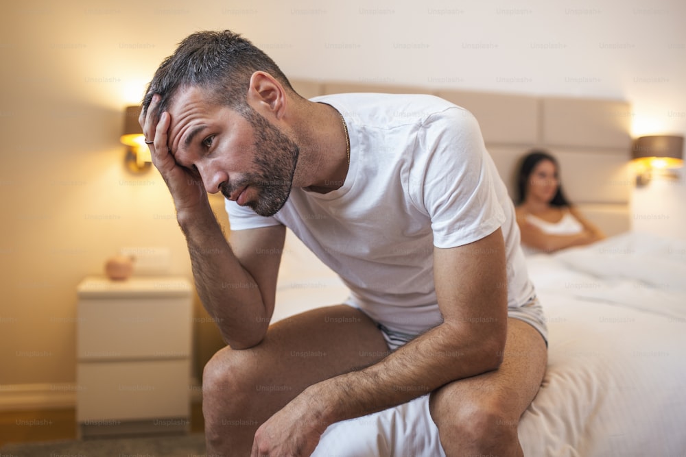 Young couple having a problem. Guy is sitting on bed and looking sadly away, his girlfriend in the background. Upset young couple having problems with sex