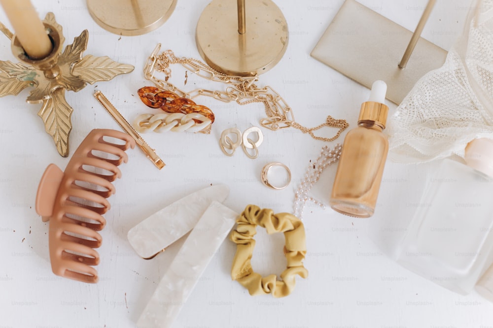 Modern summer accessories. Golden jewellery, barrettes, hair clips, cosmetics, perfume and lace lingerie on white table with vintage candles and boho mirror. Feminine essentials