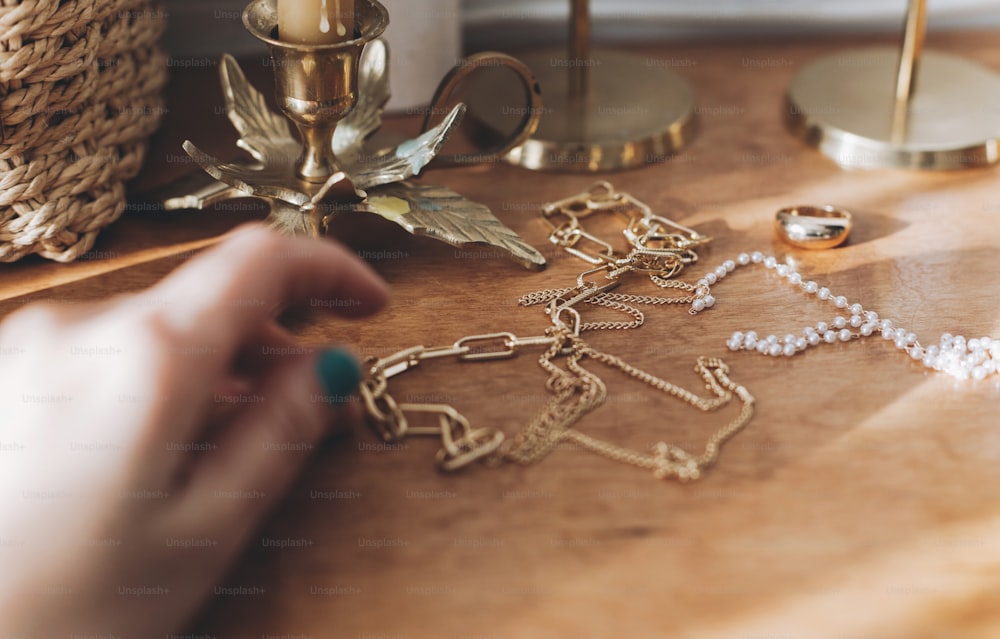 Accesorios modernos de oro y perlas y mano femenina borrosa sobre la mesa con candelabros vintage. Elegante collar dorado y anillo sobre fondo de madera a la luz del sol.