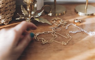 Acessórios modernos de ouro e pérola e mão feminina borrada na mesa com castiçais vintage. Colar dourado elegante e anel no fundo de madeira na luz ensolarada.