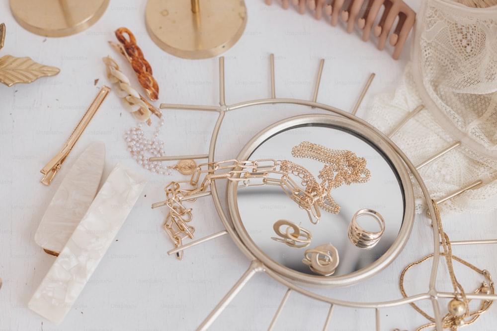 Modern summer accessories. Golden jewellery on boho mirror, barrettes, hair clips, cosmetics, perfume and lace lingerie on white table with vintage candles. Feminine essentials