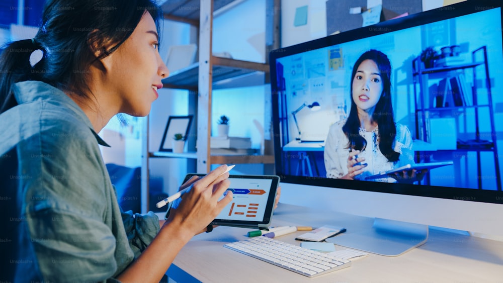 Asia businesswoman using desktop talk to colleagues about plan in video call meeting at living room. Working from house overload at night, remotely work, social distancing, quarantine for coronavirus.