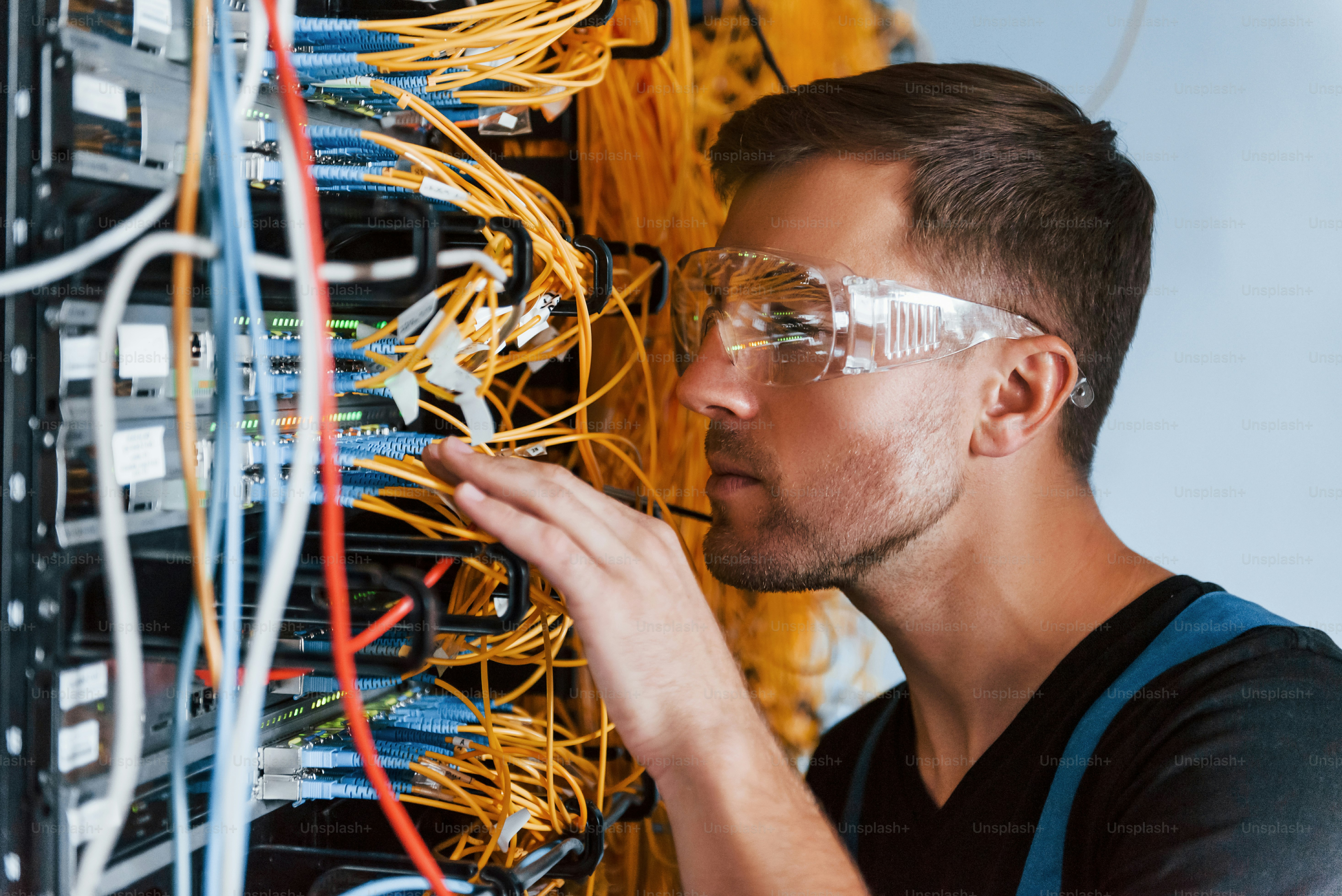 Young man in protective glasses works with internet equipment and wires in server room.