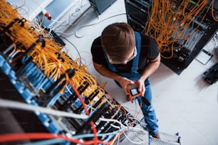 Draufsicht eines jungen Mannes in Uniform mit Messgerät, das mit Internetgeräten und Kabeln im Serverraum arbeitet.