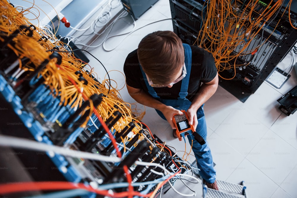 Top view of young man in uniform with measuring device that works with internet equipment and wires in server room.