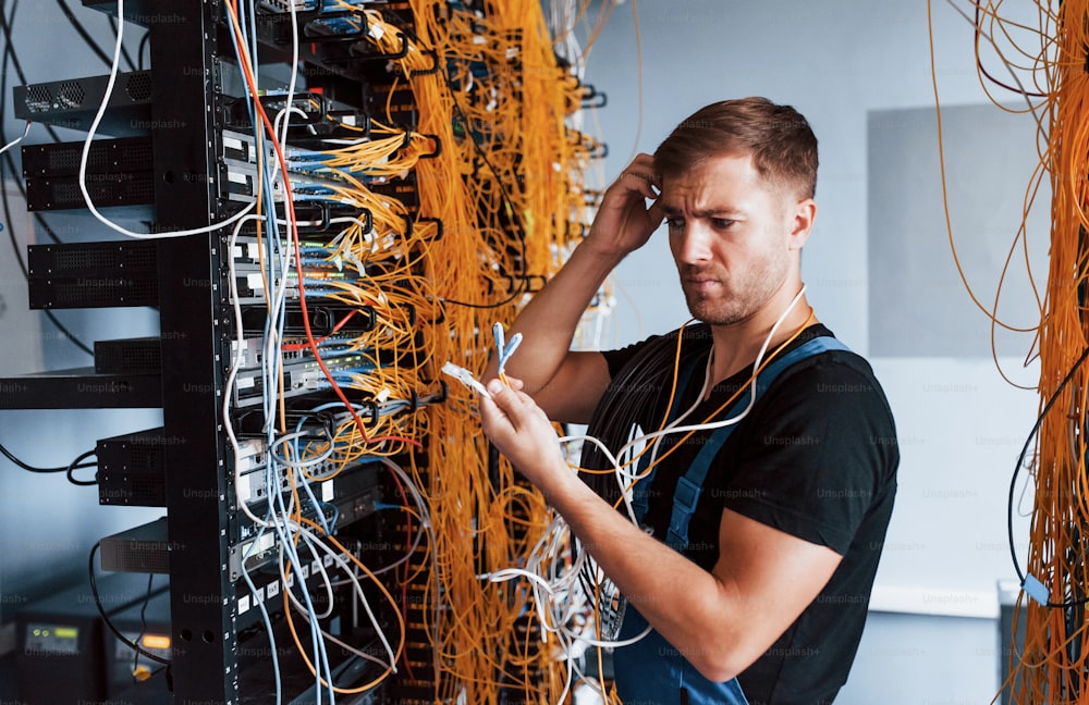 Young man in uniform feels confused and looking for a solution with internet equipment and wires in server room.