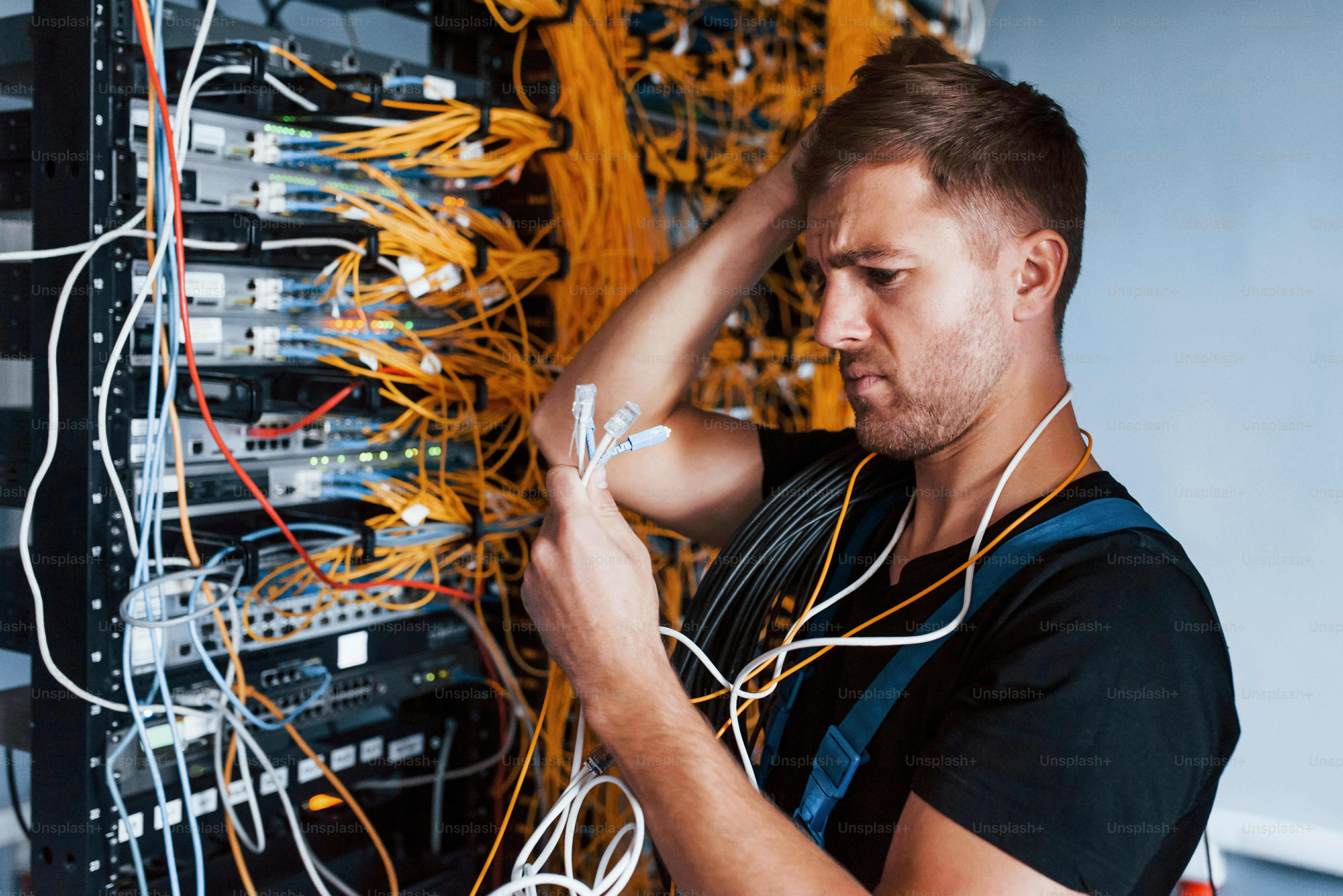 Young man in uniform feels confused and looking for a solution with internet equipment and wires in server room.