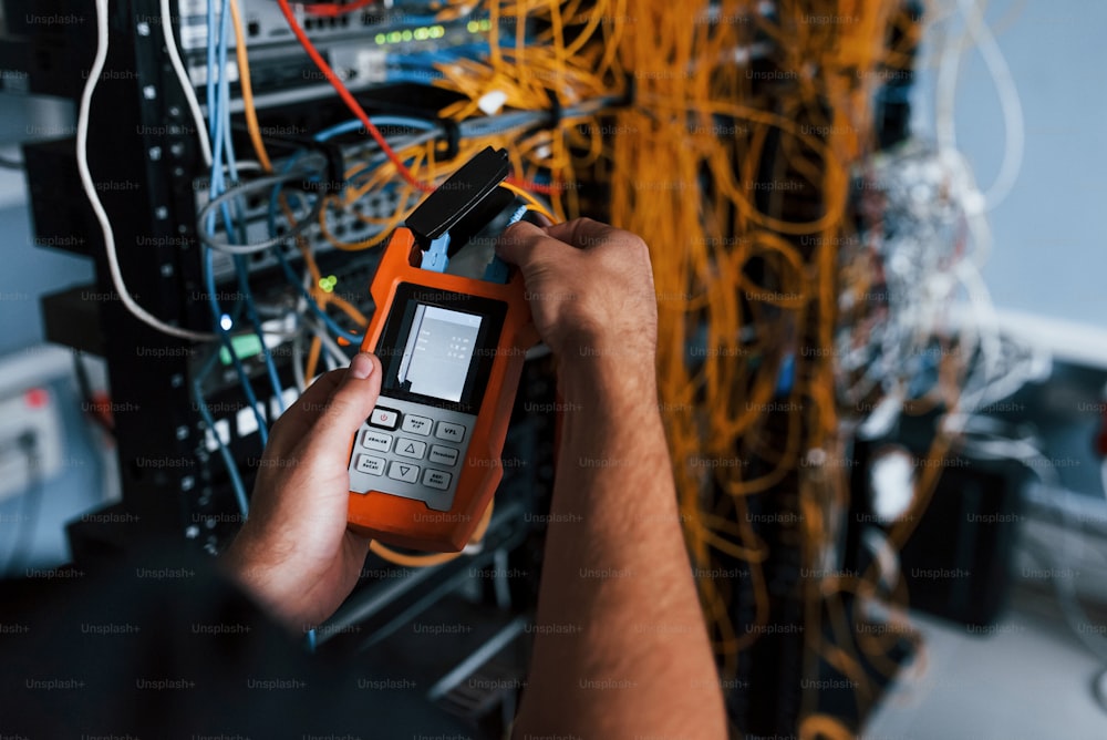 Un joven con un dispositivo de medición en las manos trabaja con equipos de Internet y cables en la sala de servidores.